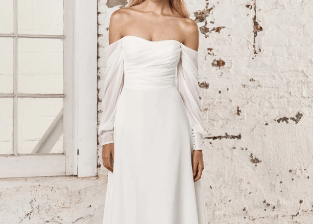 Bride wearing an off-the-shoulder wedding dress with draped sleeves and a structured bodice against a textured wall.