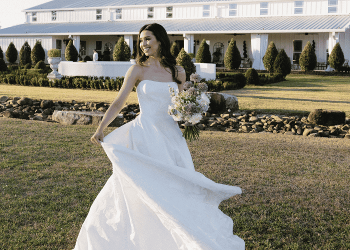 Bride wearing a strapless A-line wedding dress and holding a bouquet while standing on a lawn in front of a white event venue, with the skirt flowing as she turns.