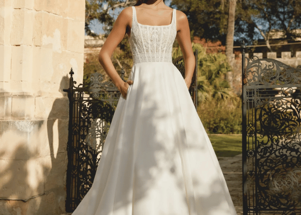 Bride in a square neckline wedding dress with lace bodice and flowing A-line skirt, standing outdoors near a garden gate.