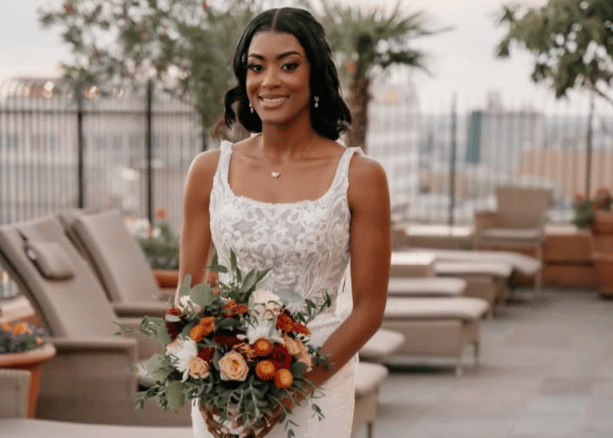 Bride holding a bouquet, wearing a scoop neckline lace wedding dress with wide straps at an outdoor venue.