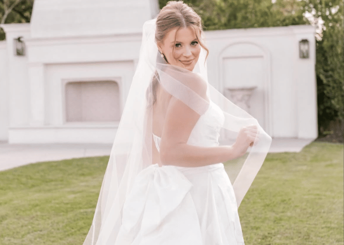 Bride in a strapless white gown and veil, smiling over her shoulder in a sunlit garden with green grass and white architectural backdrop. Elegant and joyful.