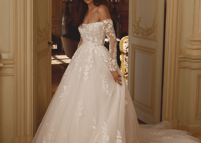 A woman stands gracefully in an ornate room, wearing an elegant, off-shoulder white wedding dress with floral lace patterns, conveying romance and sophistication.