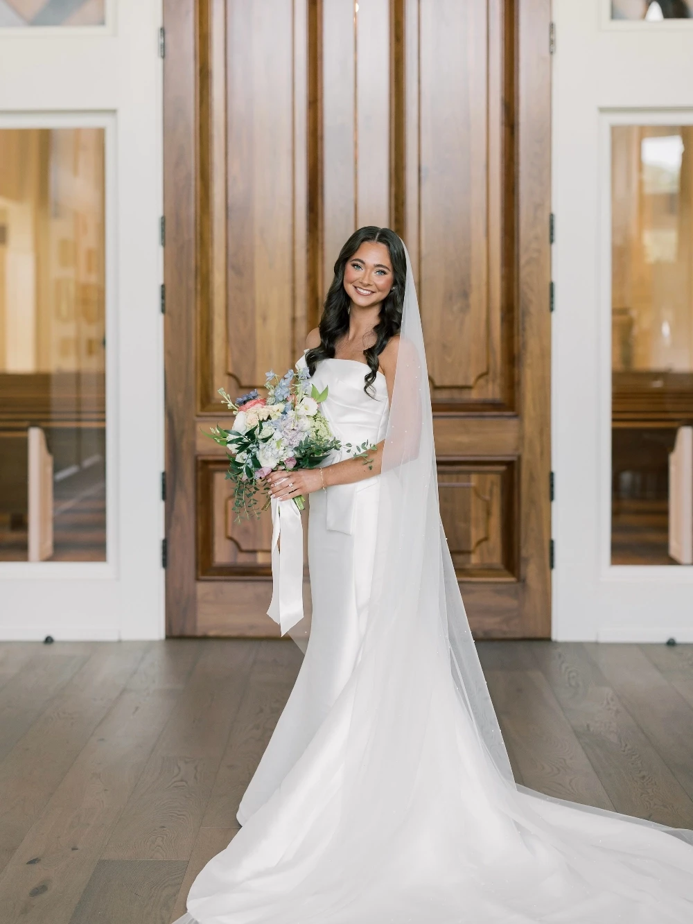 Bride in a sleek strapless wedding gown with a long train, veil, and floral bouquet.