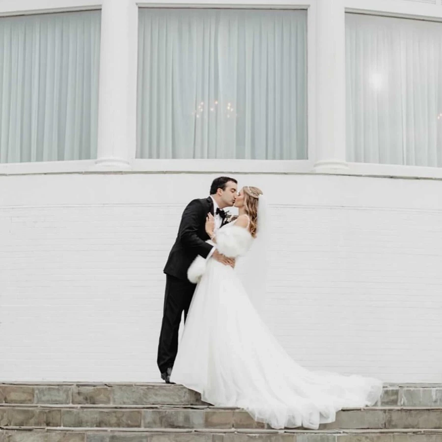 Bride and groom kissing on outdoor stone steps in front of a white building. The bride wears a long white gown with a fur wrap, and the groom is in a black tuxedo.