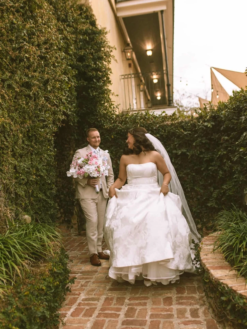 Bride in a strapless wedding gown walking with her partner through a garden courtyard.