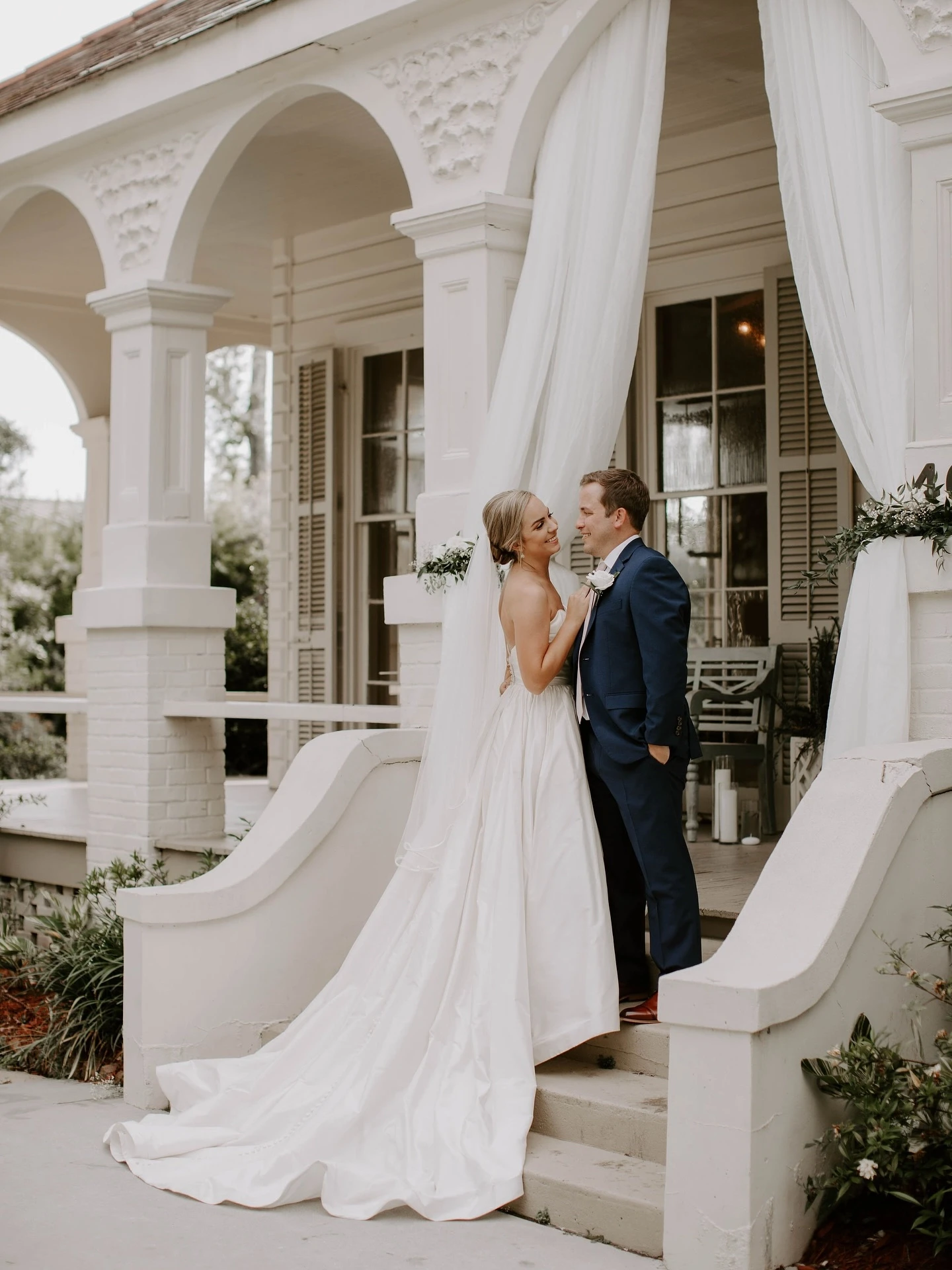 Bride and groom standing close together on the steps of a porch with white drapery. The bride wears a full satin gown with a long train, and the groom is in a navy suit.